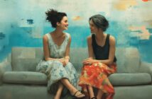 Two women sit on a gray couch, smiling at each other. One wears a light blue dress with a floral pattern, the other a dark top with a red floral skirt. The background is a bright, abstract blue and orange wall.