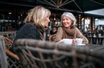 Two women sit outdoors at a café, wrapped in warm sweaters and scarves, smiling and talking while holding white coffee cups. The setting is cozy with wicker chairs and soft lighting in the background.