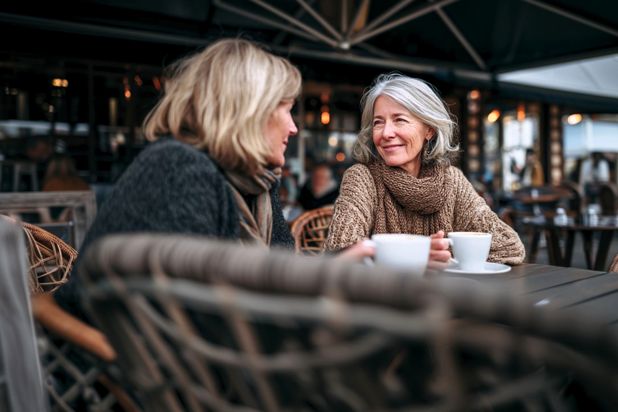 Two women sit outdoors at a café, wrapped in warm sweaters and scarves, smiling and talking while holding white coffee cups. The setting is cozy with wicker chairs and soft lighting in the background.
