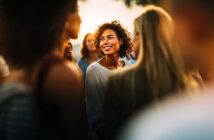 A woman with curly hair smiles brightly while standing among a group of people outdoors at sunset, creating a warm and joyful atmosphere.