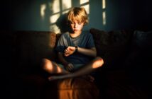 A young boy with blonde hair sits cross-legged on a couch, looking at his watch. Sunlight streams through a window, casting patterned shadows on the wall behind him.