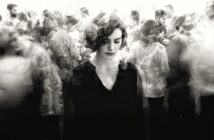 A woman with short hair stands with eyes downcast amid a blurred, bustling crowd, creating a sense of isolation and introspection despite the busy surroundings. The image is in black and white.