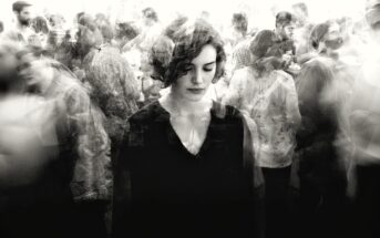 A woman with short hair stands with eyes downcast amid a blurred, bustling crowd, creating a sense of isolation and introspection despite the busy surroundings. The image is in black and white.