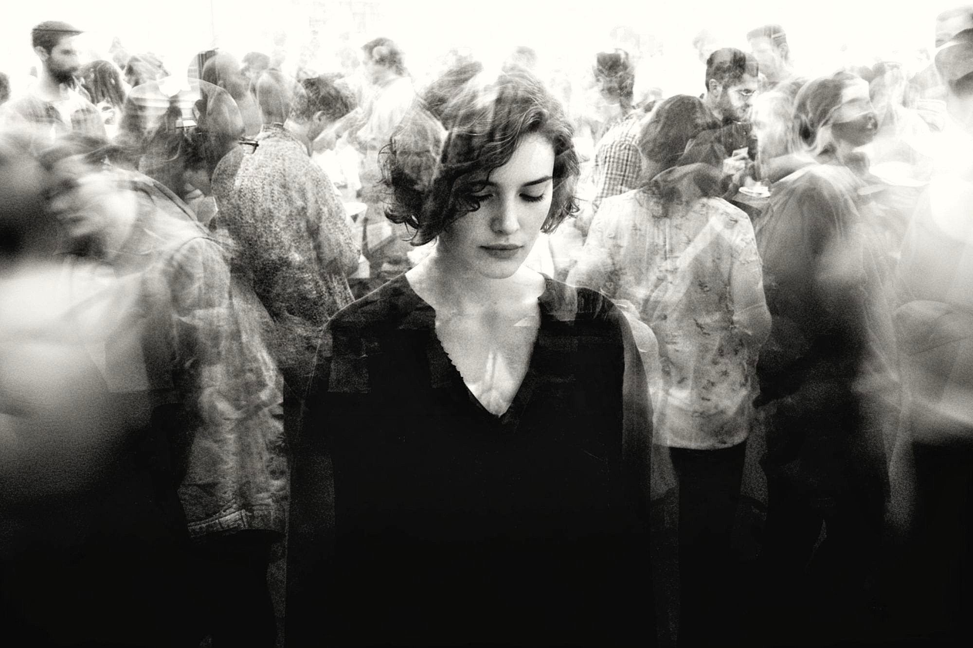 A woman with short hair stands with eyes downcast amid a blurred, bustling crowd, creating a sense of isolation and introspection despite the busy surroundings. The image is in black and white.