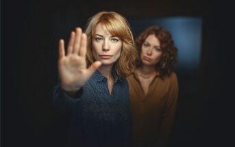 Two women stand together; the woman in front extends her hand toward the camera with a serious expression, as if signaling “stop,” while the other woman stands behind her, looking on with a neutral face.