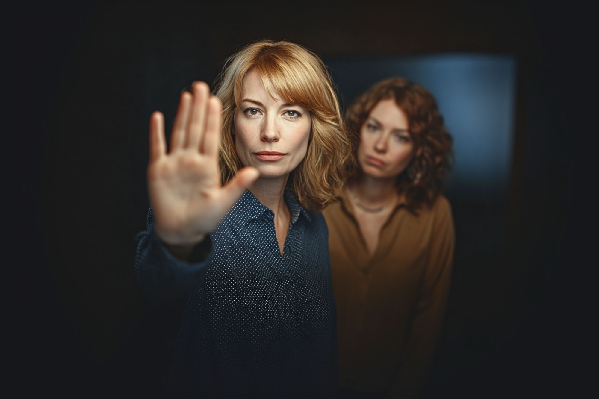 Two women stand together; the woman in front extends her hand toward the camera with a serious expression, as if signaling “stop,” while the other woman stands behind her, looking on with a neutral face.