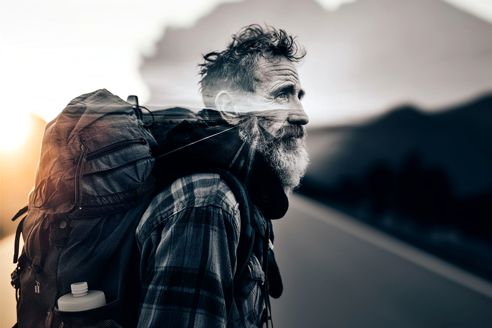 An older man with a gray beard and plaid shirt carries a large backpack, walking along a road at sunset. The background blends into a mountain landscape, giving a double exposure effect.