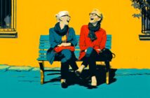 Two women sit on a blue bench against a bright yellow wall, laughing together. Both wear scarves and glasses; one is in a blue coat, the other in a red coat. Sunlight casts shadows, creating a cheerful outdoor scene.