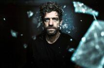 A man with curly hair and a beard stares intently ahead as shards of glass appear to shatter and fly around him against a dark background, creating a dramatic and intense atmosphere.