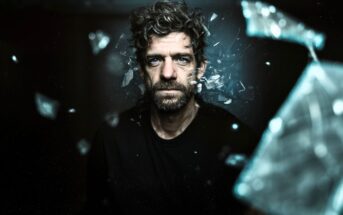 A man with curly hair and a beard stares intently ahead as shards of glass appear to shatter and fly around him against a dark background, creating a dramatic and intense atmosphere.