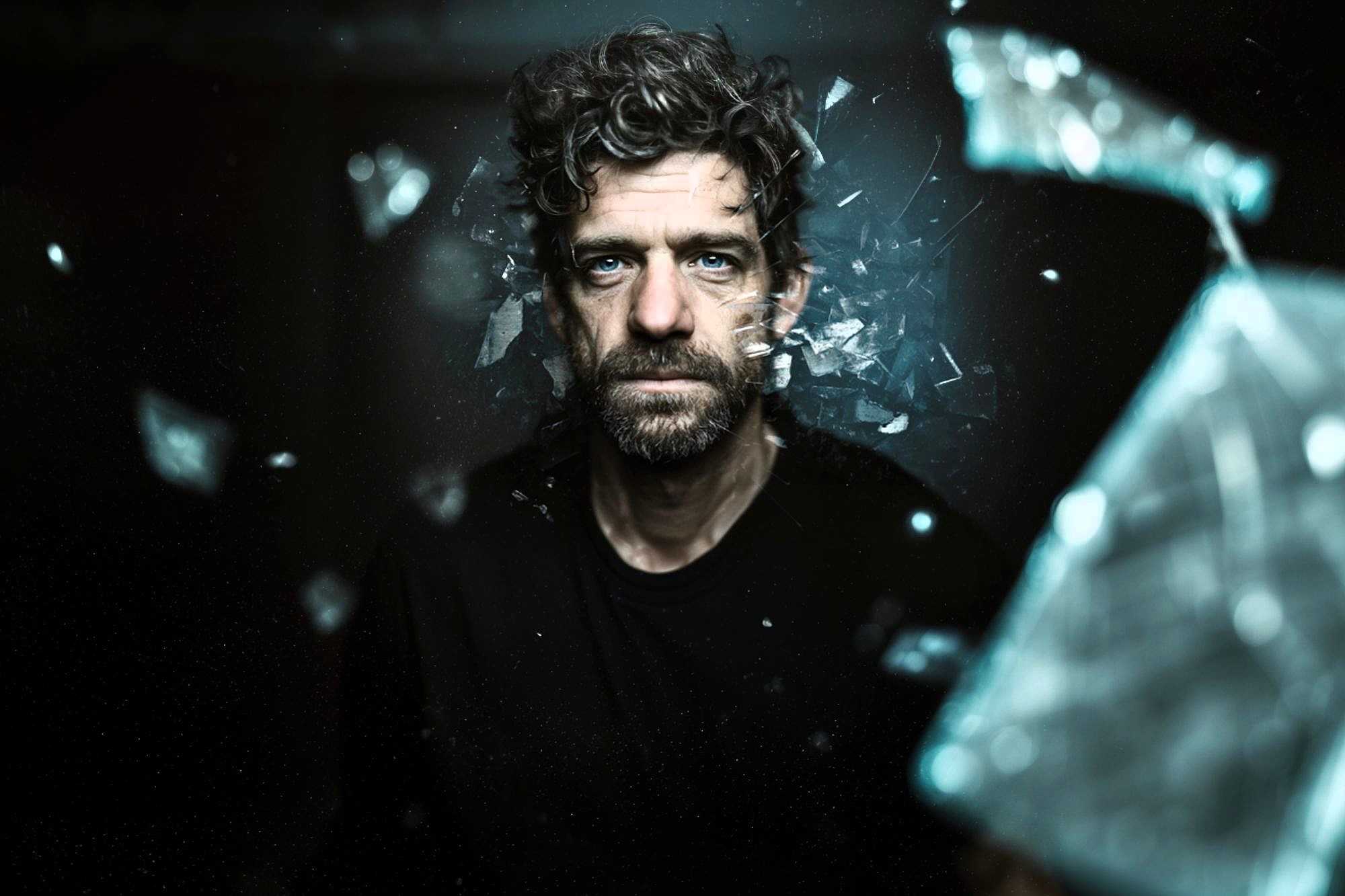 A man with curly hair and a beard stares intently ahead as shards of glass appear to shatter and fly around him against a dark background, creating a dramatic and intense atmosphere.