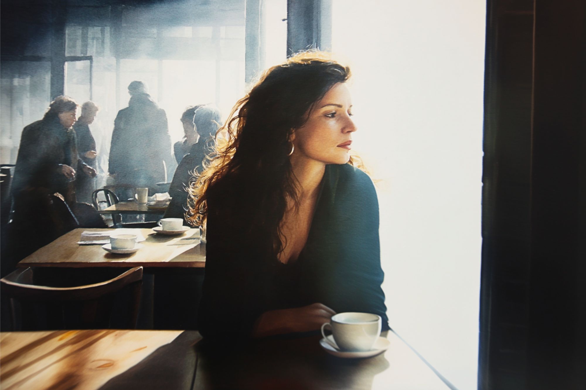 A woman with wavy hair sits alone at a café table with a cup of coffee, gazing out a sunlit window. Other people are seen in the background, creating a calm, contemplative atmosphere.
