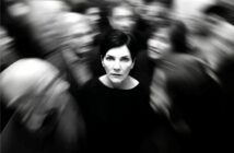 A woman with short dark hair stands still and looks ahead, sharply focused amid a blurred crowd moving around her, suggesting isolation or feeling overwhelmed. The photo is in black and white.