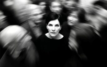 A woman with short dark hair stands still and looks ahead, sharply focused amid a blurred crowd moving around her, suggesting isolation or feeling overwhelmed. The photo is in black and white.
