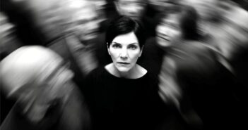 A woman with short dark hair stands still and looks ahead, sharply focused amid a blurred crowd moving around her, suggesting isolation or feeling overwhelmed. The photo is in black and white.
