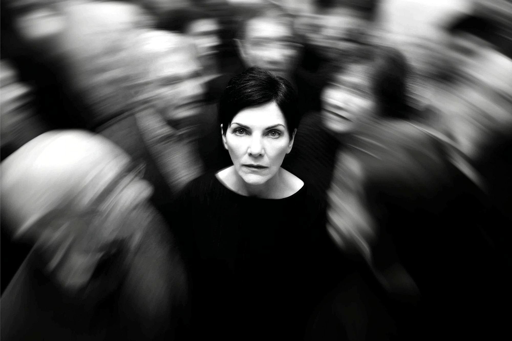 A woman with short dark hair stands still and looks ahead, sharply focused amid a blurred crowd moving around her, suggesting isolation or feeling overwhelmed. The photo is in black and white.
