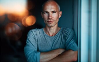 A man with short hair and a trimmed beard stands indoors with arms crossed, wearing a gray shirt. Warm lights are blurred in the background, creating a soft, relaxed atmosphere.