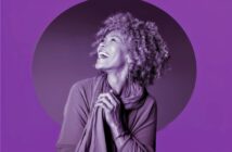 A joyful older woman with curly hair smiles and looks upward, hands clasped in front of her, against a purple background with a darker circle behind her.