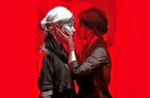 Two women stand close together against a vivid red background. One gently holds the other's face with both hands, their expressions intense and intimate, suggesting a deep emotional connection between them.