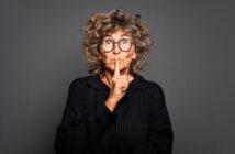 Older woman with curly hair and glasses wearing a black sweater, holding her finger to her lips in a "shh" gesture, standing against a plain dark gray background.