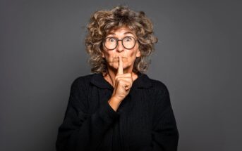 Older woman with curly hair and glasses wearing a black sweater, holding her finger to her lips in a "shh" gesture, standing against a plain dark gray background.
