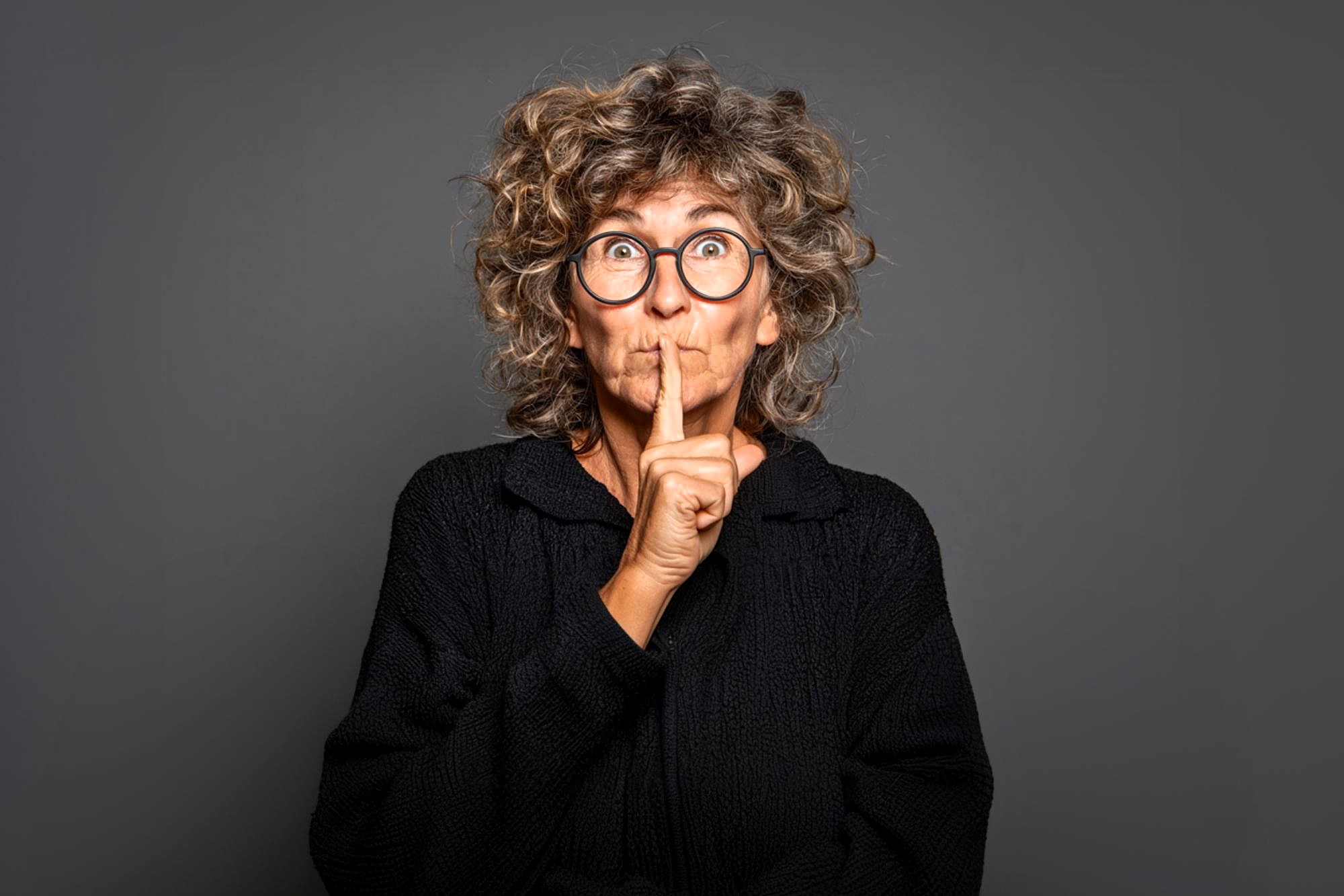 Older woman with curly hair and glasses wearing a black sweater, holding her finger to her lips in a "shh" gesture, standing against a plain dark gray background.
