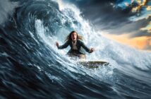 A person with long hair, wearing a jacket and pants, looks surprised while surfing a large, curling wave under a dramatic cloudy sky at sunset.