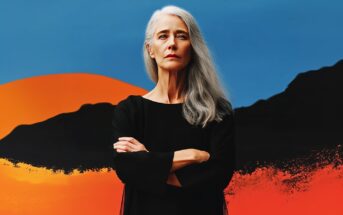 A woman with long gray hair and a serious expression stands with arms crossed, wearing a black top, in front of a vibrant blue, orange, and black abstract background.