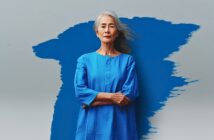 An older woman with long gray hair stands confidently with arms crossed, wearing a bright blue dress. She is posed in front of a gray background featuring a bold, abstract blue paint streak.
