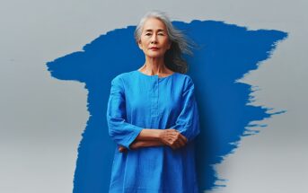 An older woman with long gray hair stands confidently with arms crossed, wearing a bright blue dress. She is posed in front of a gray background featuring a bold, abstract blue paint streak.