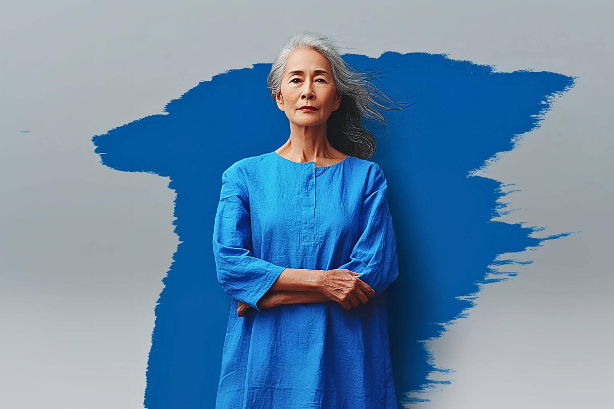 An older woman with long gray hair stands confidently with arms crossed, wearing a bright blue dress. She is posed in front of a gray background featuring a bold, abstract blue paint streak.