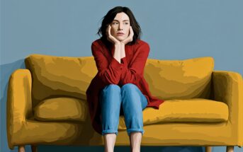A woman with dark hair, wearing a red shirt and jeans, sits barefoot on a yellow couch with her chin resting on her hands, looking thoughtful against a plain blue wall.