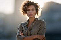 A woman with curly hair stands outdoors, wearing a loose, buttoned shirt with her arms crossed, looking confidently at the camera against a softly blurred background and sunlight.