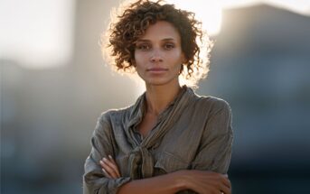 A woman with curly hair stands outdoors, wearing a loose, buttoned shirt with her arms crossed, looking confidently at the camera against a softly blurred background and sunlight.