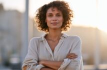 A woman with curly hair stands with her arms crossed, smiling at the camera during sunset.