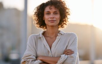 A woman with curly hair stands with her arms crossed, smiling at the camera during sunset.