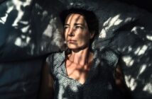A woman lies on a bed, looking upward with a thoughtful expression. Dappled sunlight and shadows from leaves create patterns across her face, pillow, and shirt.