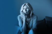 A thoughtful middle-aged woman with light hair sits indoors, resting her chin on her hand and gazing into the distance. The image has a blue tint, creating a calm and contemplative mood.
