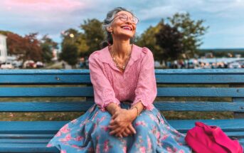 An older woman with gray hair and glasses, wearing a pink blouse and floral skirt, sits smiling on a blue bench outdoors. A bright pink bag rests beside her, with trees and a cloudy sky in the background.
