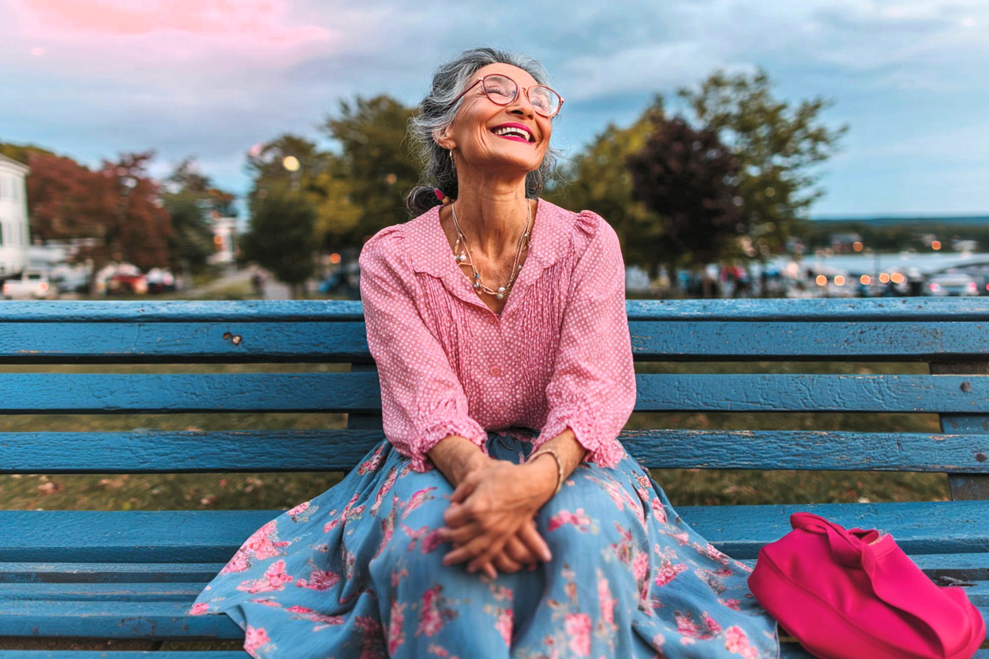 An older woman with gray hair and glasses, wearing a pink blouse and floral skirt, sits smiling on a blue bench outdoors. A bright pink bag rests beside her, with trees and a cloudy sky in the background.