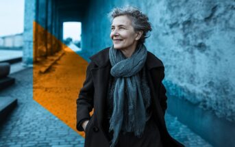 An older woman with curly gray hair, wearing a coat and scarf, smiles while walking on a cobblestone path. A translucent orange geometric shape overlays part of the image, contrasting with the background.