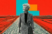 An older woman in a black-and-white houndstooth coat stands on a patterned path in a surreal, vividly colored landscape with bold red ground and geometric shapes in blue and yellow against a red sky.