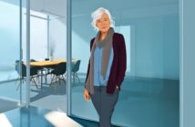 An older woman with white hair, wearing a scarf, cardigan, and blouse, stands smiling in front of a glass-walled office conference room with a table and chairs.