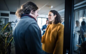 A woman with curly hair in a mustard sweater smiles at another person from across a room. Other people are visible in the background, including a reflection in a glass door.