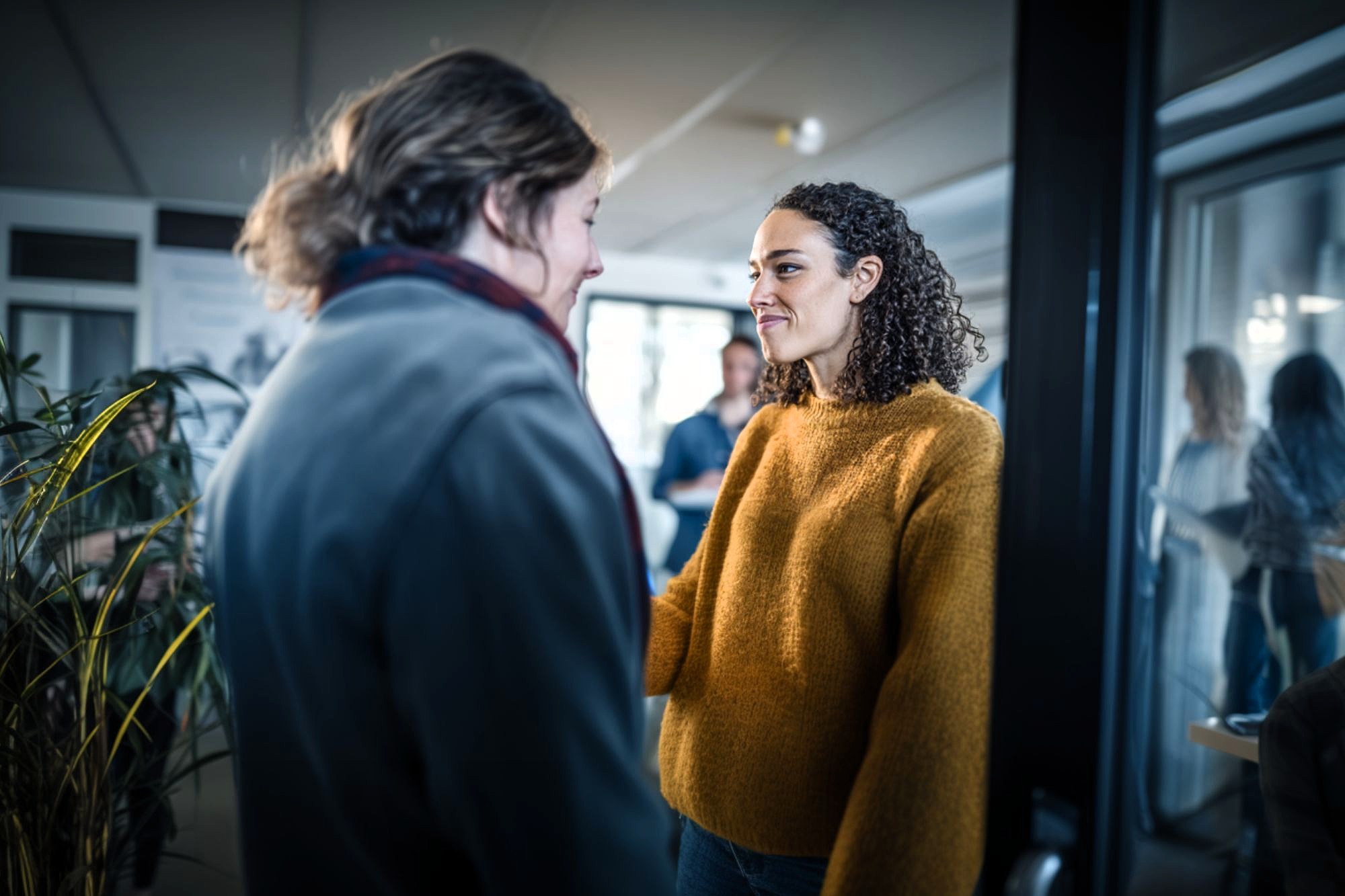 A woman with curly hair in a mustard sweater smiles at another person from across a room. Other people are visible in the background, including a reflection in a glass door.