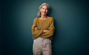 Smiling older woman with gray hair in a ponytail, wearing a mustard sweater and beige pants, stands with arms crossed against a dark green background.
