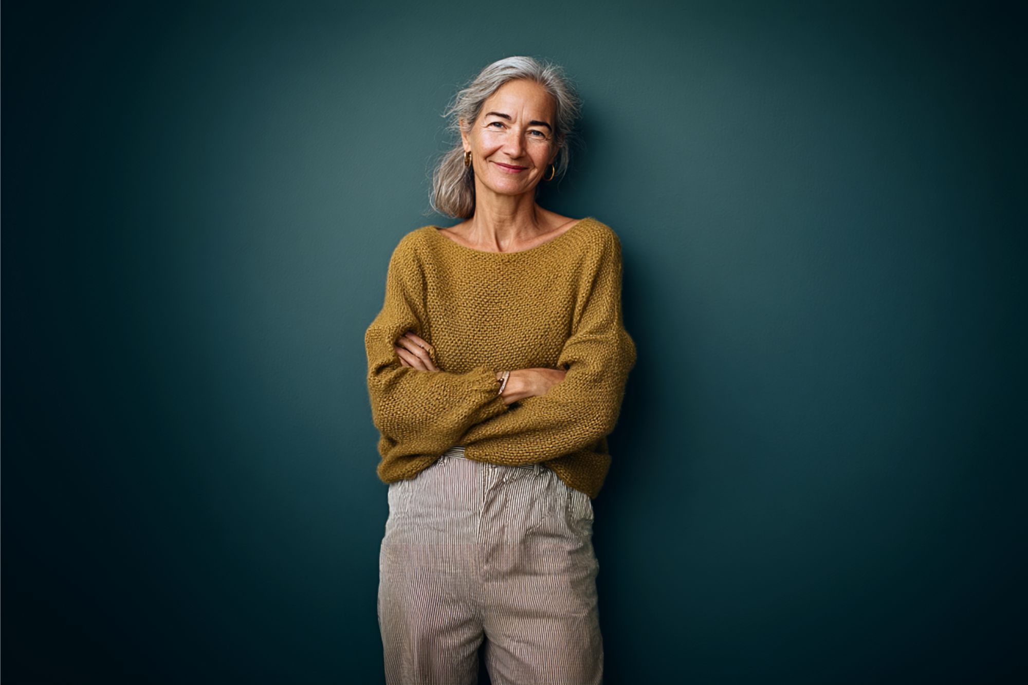 Smiling older woman with gray hair in a ponytail, wearing a mustard sweater and beige pants, stands with arms crossed against a dark green background.