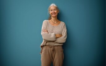 An older woman with long gray hair smiles while standing in front of a teal wall. She is wearing a loose beige sweater and light brown pants, with her arms crossed.