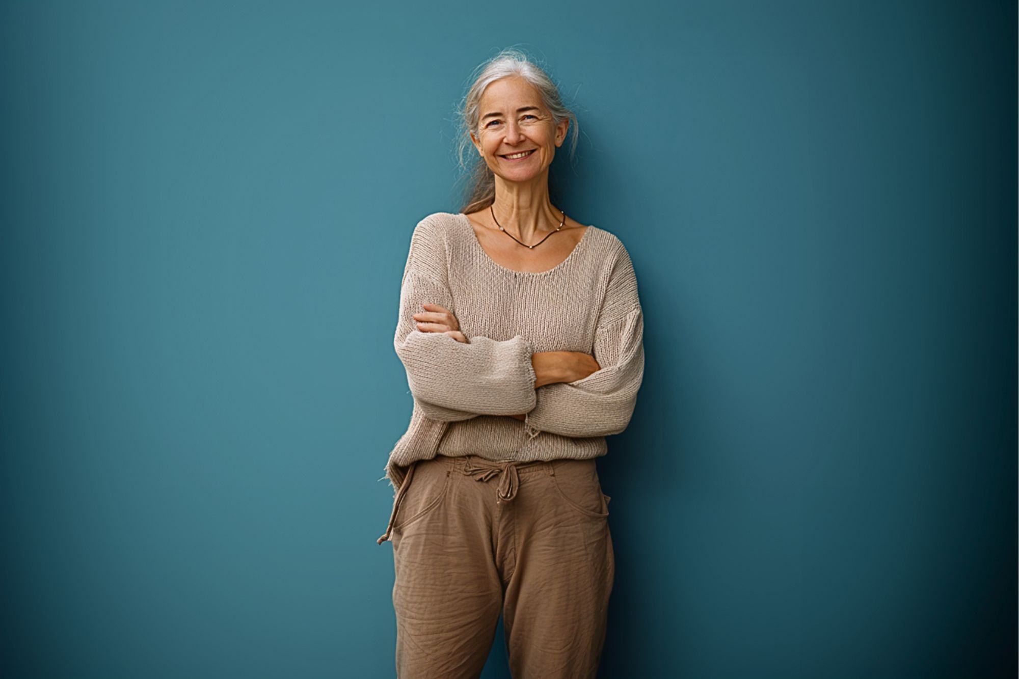 An older woman with long gray hair smiles while standing in front of a teal wall. She is wearing a loose beige sweater and light brown pants, with her arms crossed.