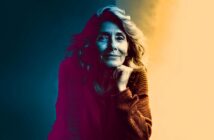 A woman with wavy hair and a warm sweater rests her chin on her hand, smiling softly. The image uses dramatic lighting with blue tones on one side and warm yellow on the other, creating a vivid color contrast.